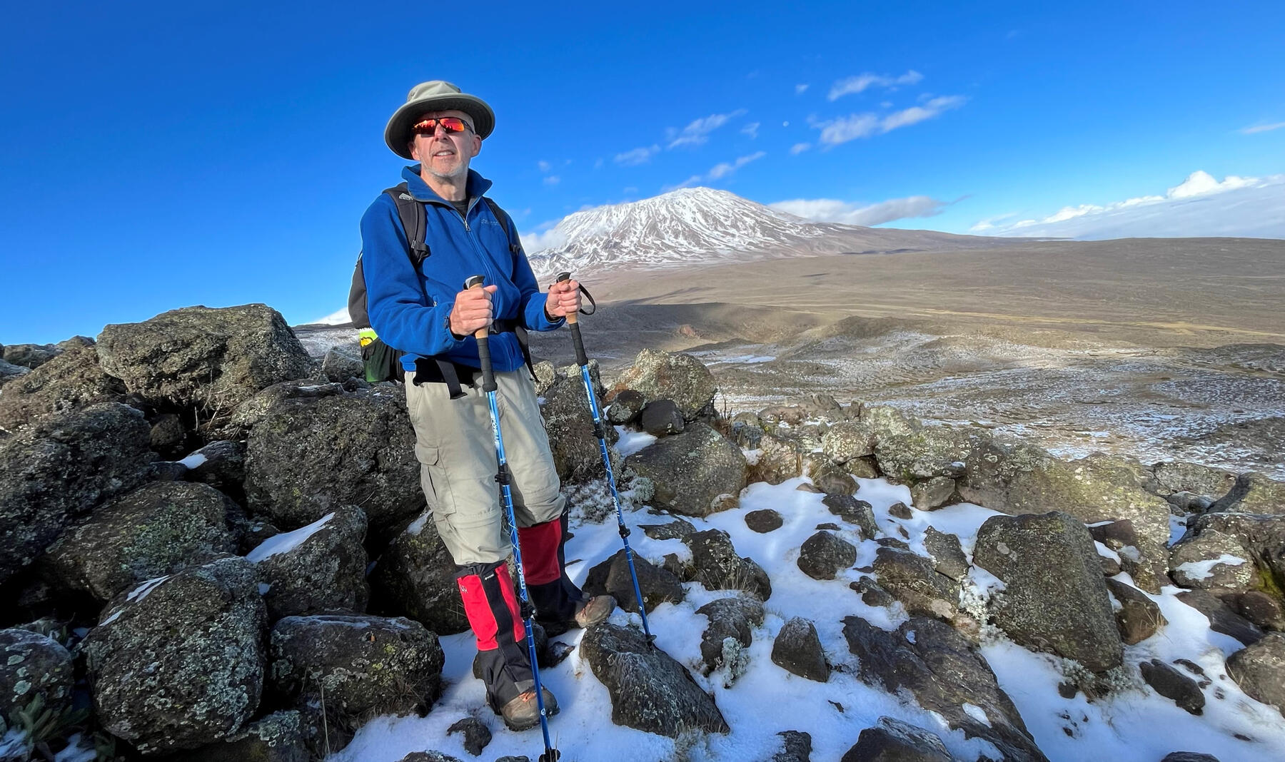 Mike hiking among volcanic rocks with the snow-covered summit of Mount Kilimanjaro behind him.